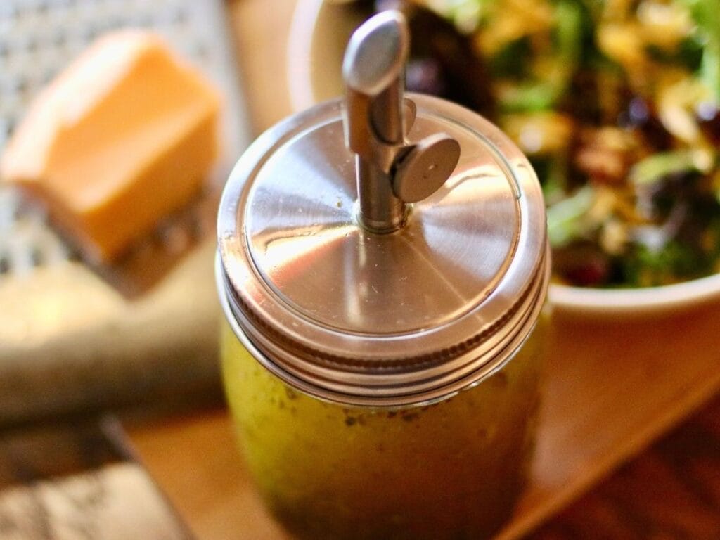 Jar of Italian vinaigrette with pour spout with salad and a block of cheese on a grater