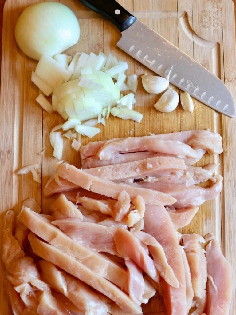 Chopped onion, garlic cloves, and sliced chicken on a cutting board.