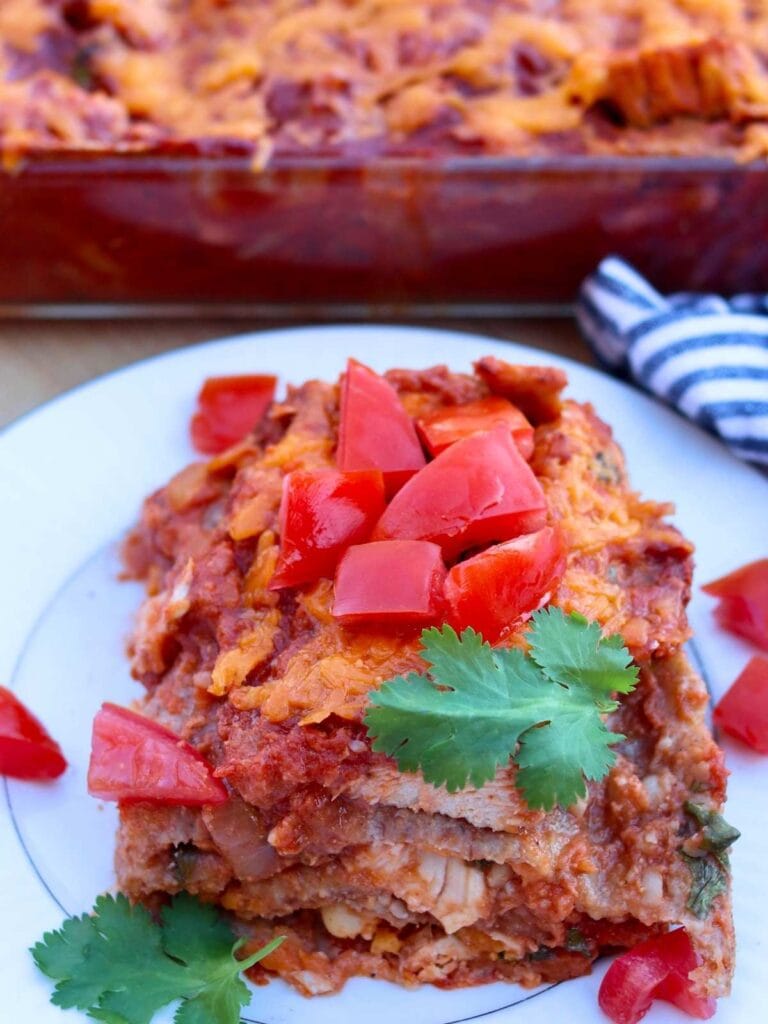 Slice of chicken enchilada casserole on a plate with chopped tomatoes and fresh cilantro.