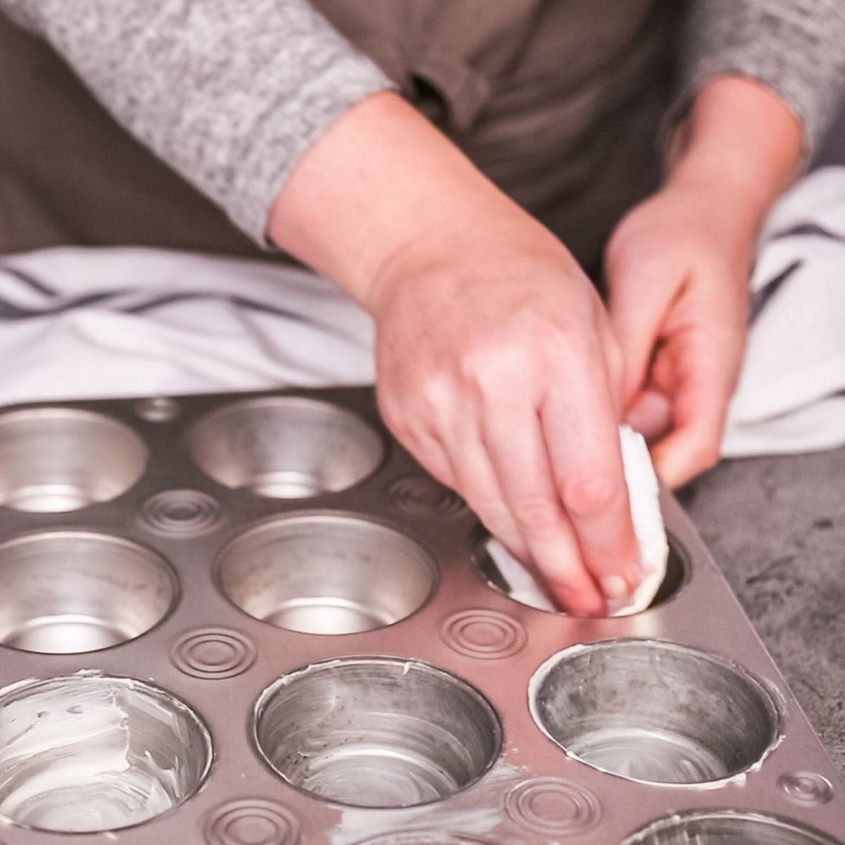 Hands using a paper towel to grease a muffin pan with butter.