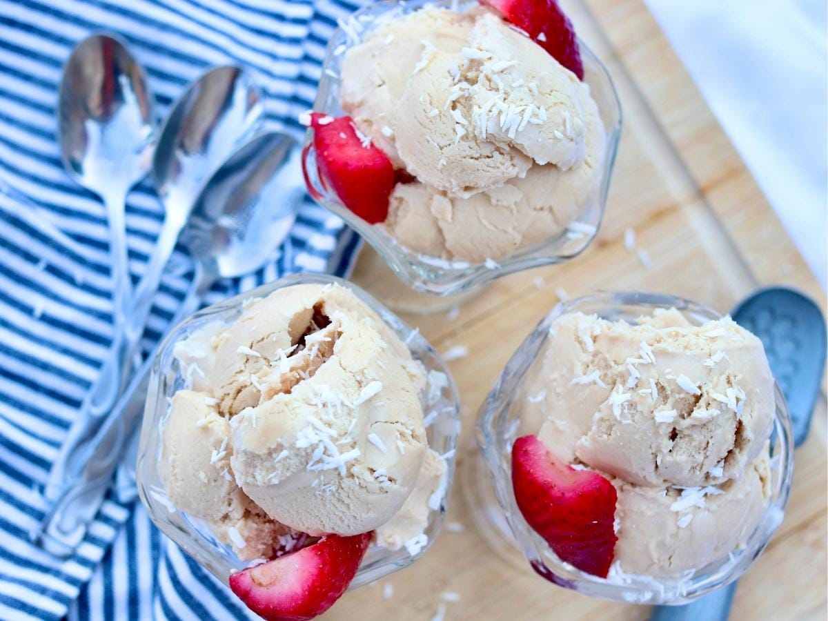 Three spoons and three bowls of ice cream topped with strawberries and coconut.