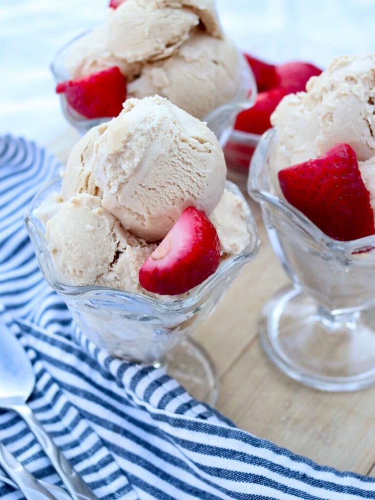 Three glass ice cream bowls filled with ice cream and garnished with strawberries beside spoons.