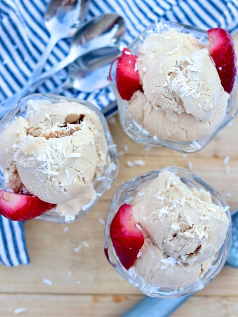 Top view of three glass ice cream bowls filled with ice cream and garnished with coconut and strawberries and three spoons on a napkin.