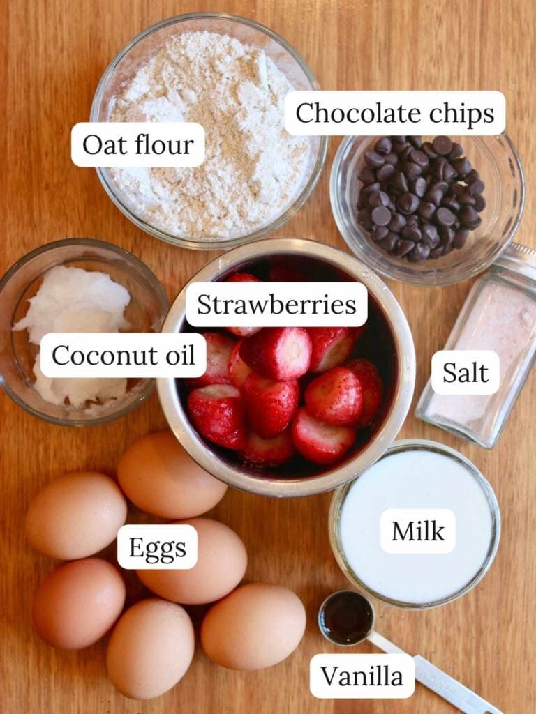 Ingredients for strawberry Dutch baby including strawberries, eggs, oat flour, and more on a wood background.