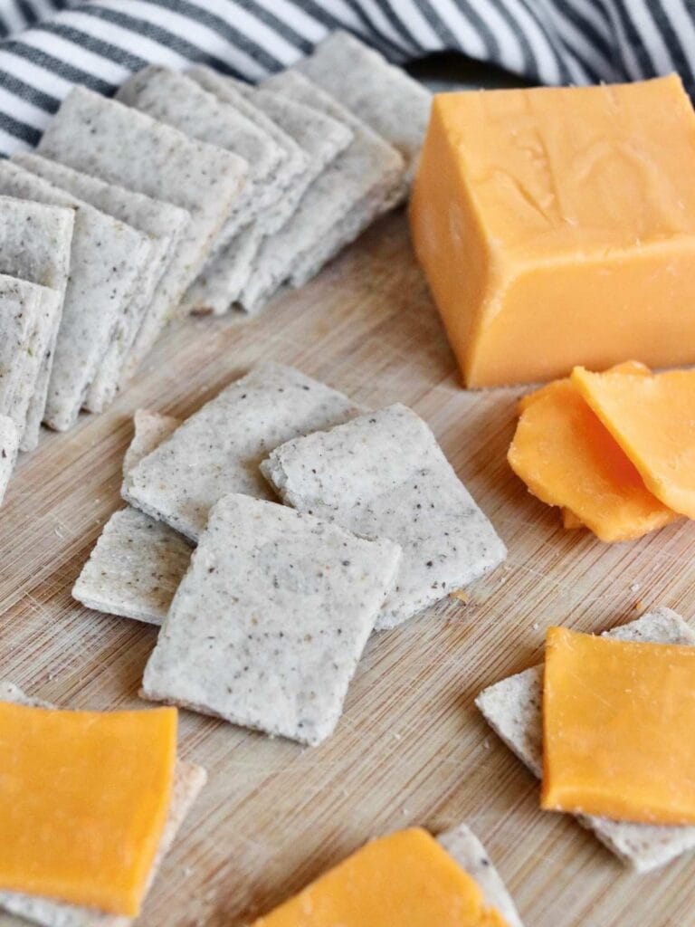 Pile of almond flour crackers with a block of cheese and slices of cheese on individual crackers.