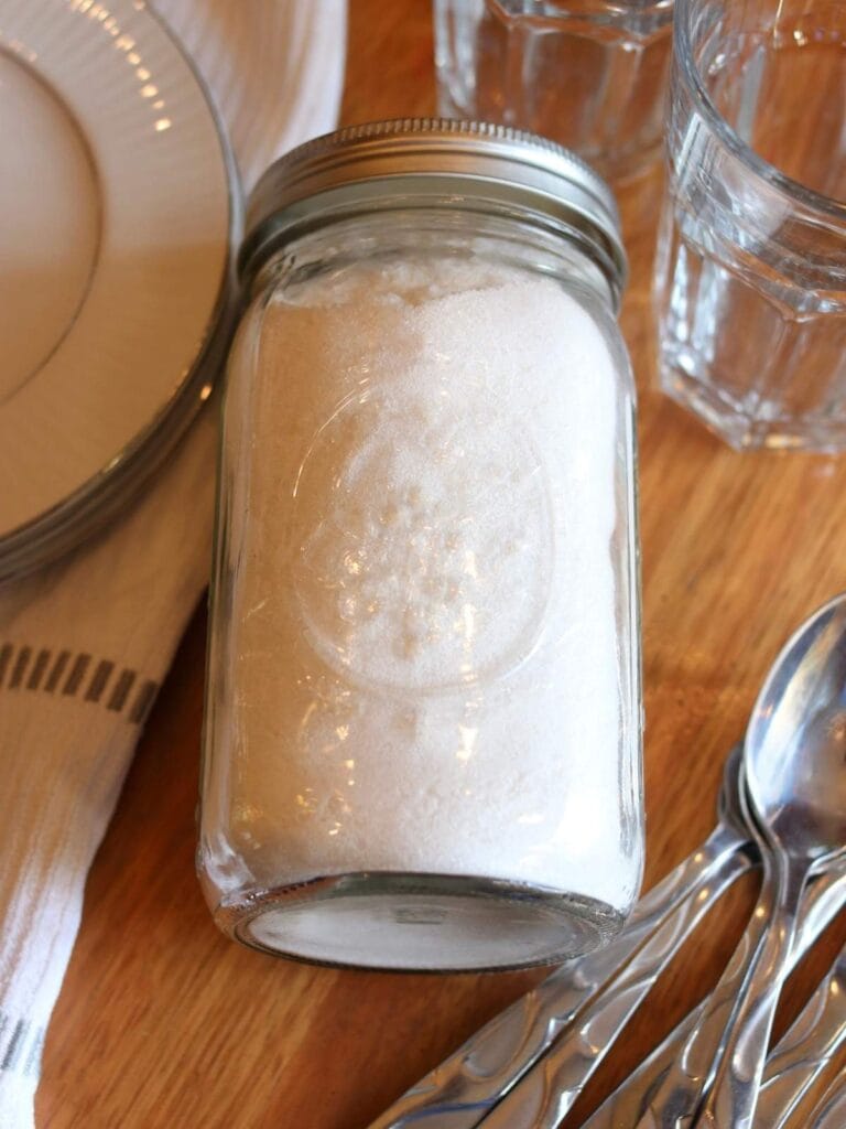 Mason jar with homemade dishwasher detergent with clean plates, spoons, and glasses.