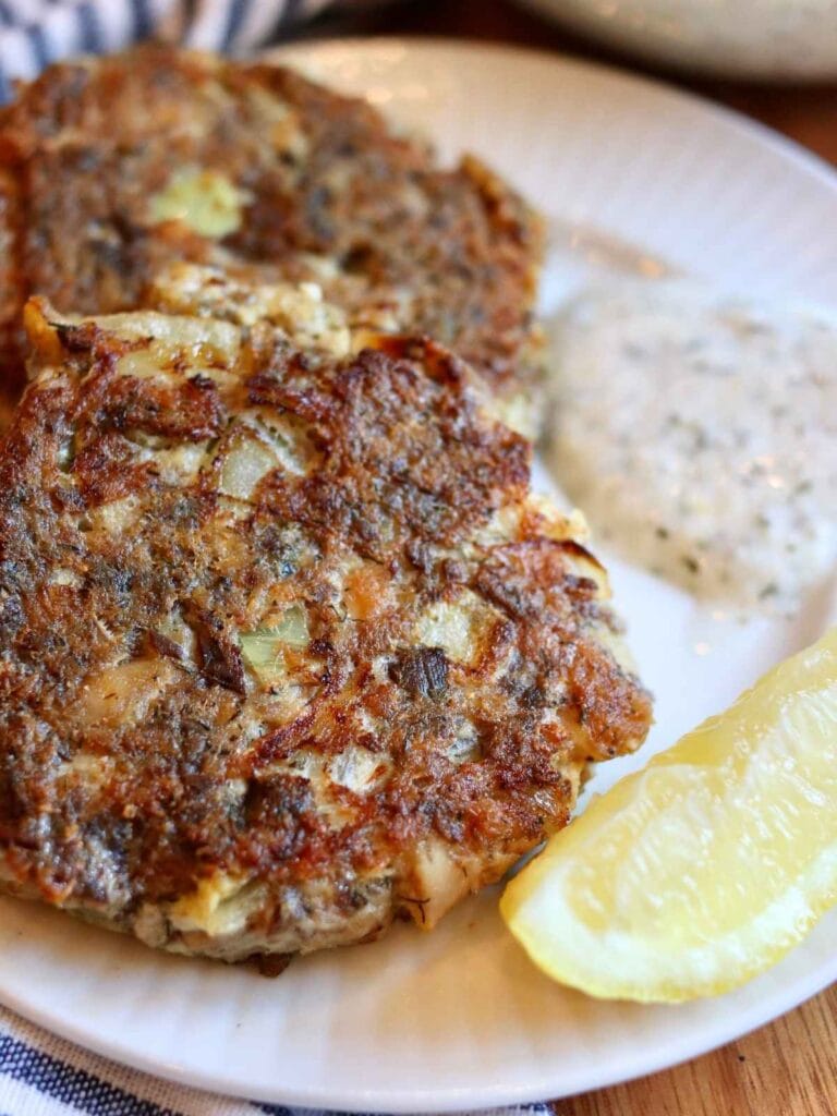 2 sardine fish cakes on a plate with yogurt dill sauce and a lemon slice.