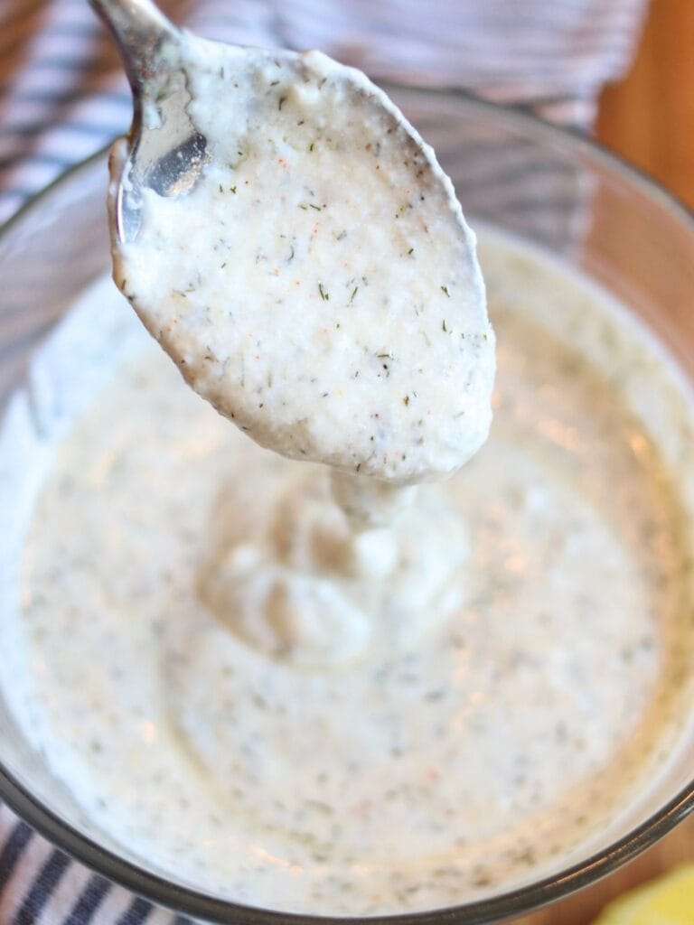 Yogurt dill sauce pouring off of a spoon into a bowl of the sauce.