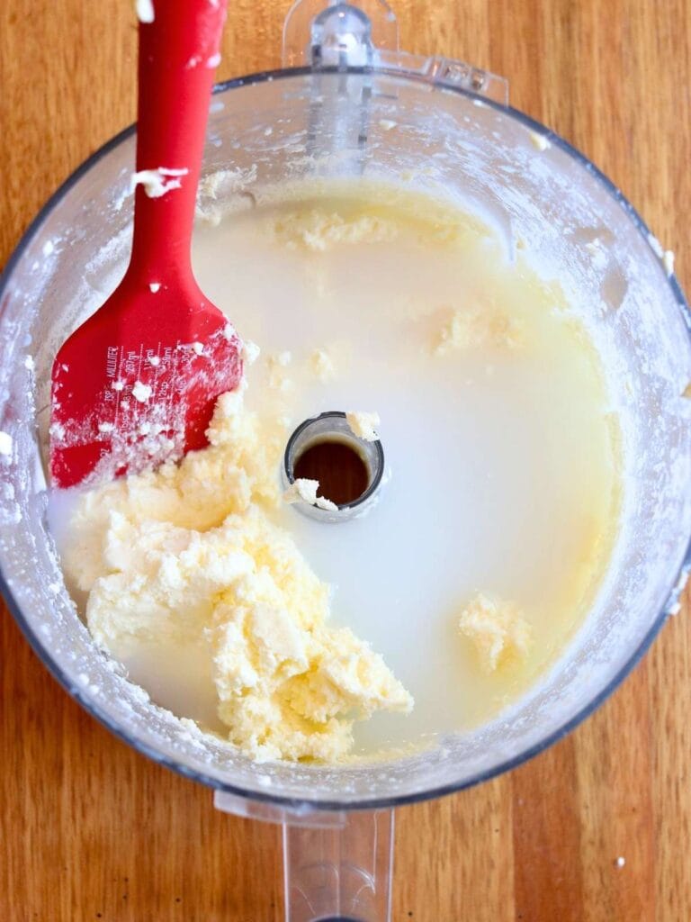Rinsing buttermilk from freshly blended homemade butter in a food processor bowl with a red spatula.