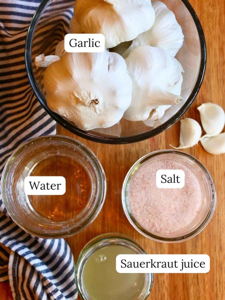Ingredients for fermented garlic including garlic, water, salt, and sauerkraut juice on a wooden surface.