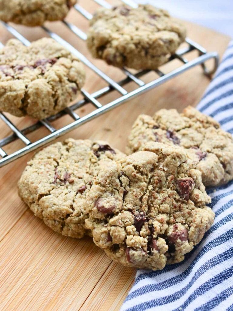 Three oat flour chocolate chip cookies beside a cooling rack with more cookies.
