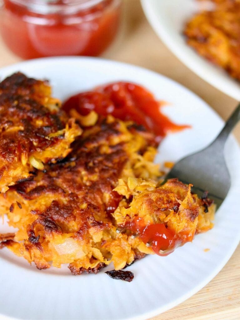 A bite of a sweet potato fritter with ketchup on a fork resting on a plate with two golden crispy fritters.