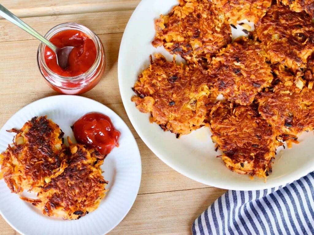 Freshly made sweet potato fritters served on a white plate with a side of ketchup on a wooden table.