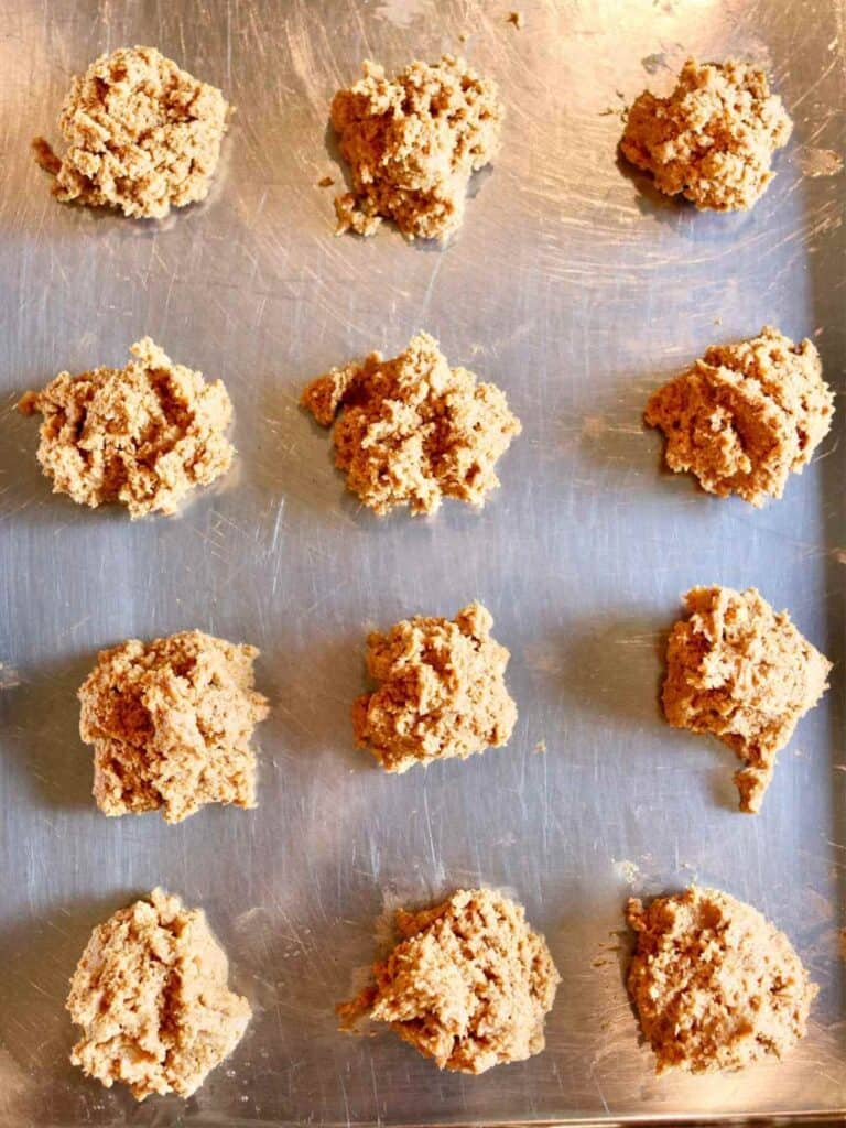 Twelve evenly spaced cookie dough scoops on a metal baking sheet, ready for the oven. The dough is golden brown and has a soft texture.