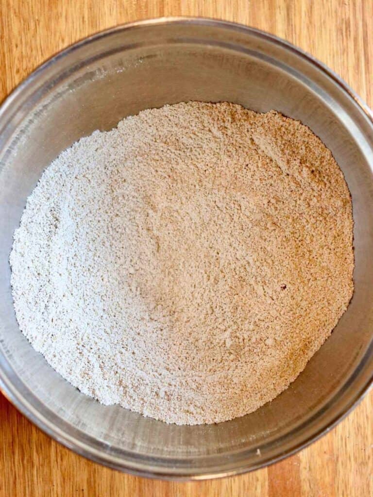 A metal bowl filled with oat flour and other dry ingredients sits on a wooden surface. The texture is fine and even, suggesting freshly mixed ready for baking.