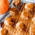 Gluten free pumpkin cookies with rich maple glaze are cooling on a wire rack on a wooden table. A large pumpkin and striped cloth add an autumnal touch.