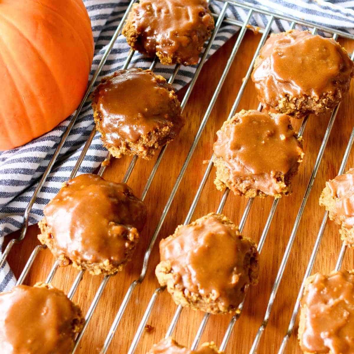 Gluten free pumpkin cookies with rich maple glaze are cooling on a wire rack on a wooden table. A large pumpkin and striped cloth add an autumnal touch.