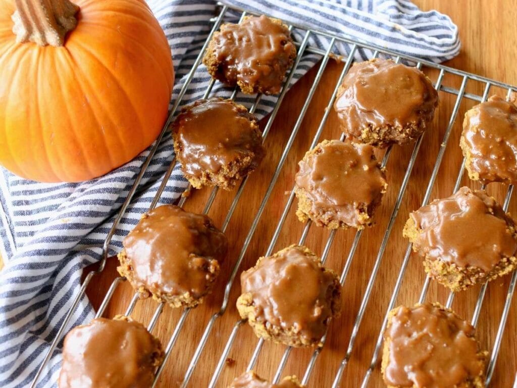 Gluten free pumpkin cookies with shiny maple glaze are cooling on a wire rack over a wooden surface. A large pumpkin and striped cloth add an autumnal touch.