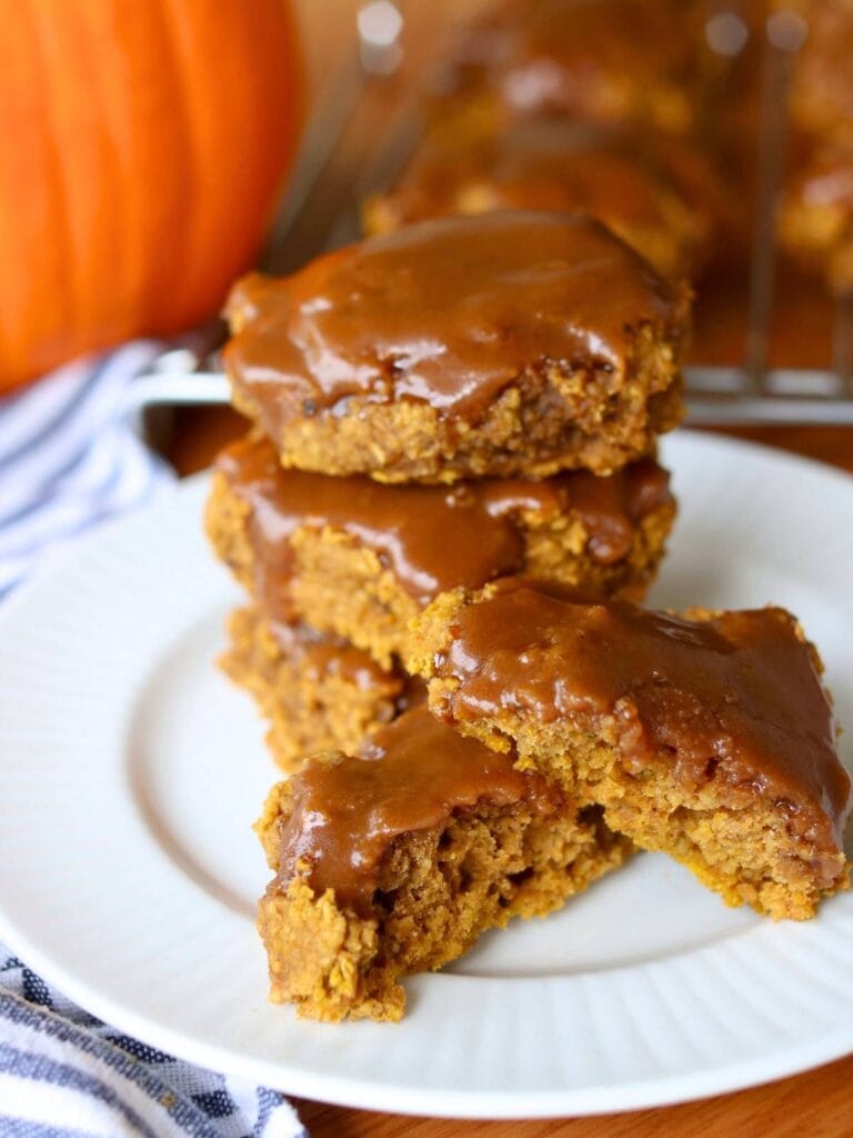A stack of gluten free pumpkin cookies with maple glaze on a white plate, sitting on a wooden table. A whole pumpkin and striped cloth are in the background, evoking autumn vibes.