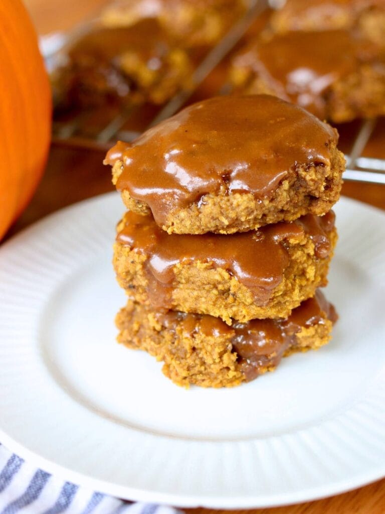 Stack of three gluten free pumpkin cookies on a white plate. The cookies are thick, moist, and topped with a shiny maple glaze. A warm, cozy autumn treat.