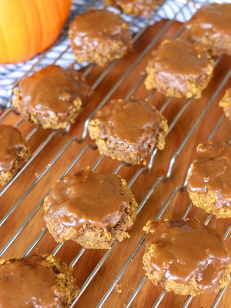 Gluten free pumpkin cookies with a glossy maple glaze cool on a wire rack. A pumpkin is in the background, hinting at fall flavors and setting a cozy tone.