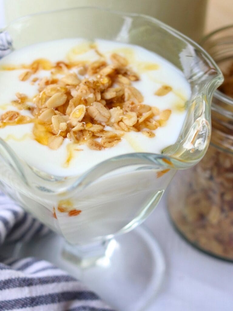 Glass dish of goat yogurt topped with granola and honey drizzle on a white surface. A striped cloth and jar of granola are visible in the background.