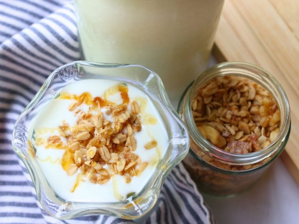 A glass bowl of goat yogurt topped with granola and honey sits beside a jar of granola on a striped cloth, conveying a fresh and healthy breakfast vibe.