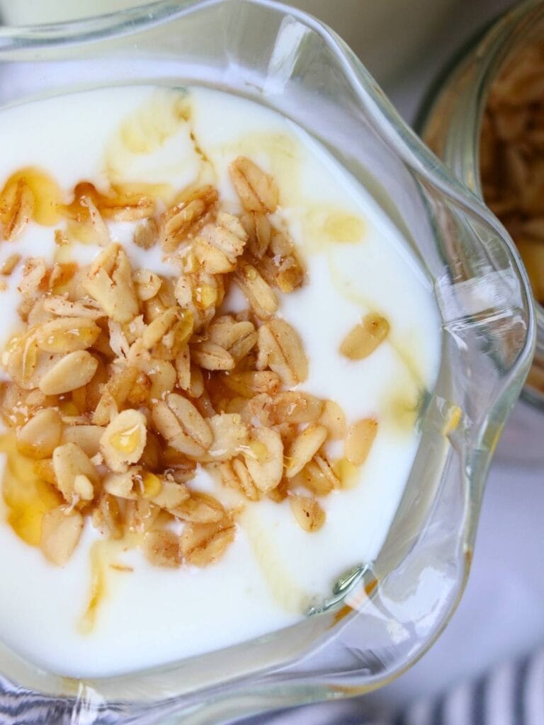 A glass cup of creamy goat yogurt is topped with golden granola drizzled with honey. The texture of the oats stands out against the smooth homemade yogurt.
