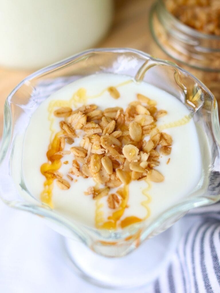 A glass dish of creamy goat yogurt topped with granola and drizzled with honey. The setting is bright and inviting, evoking a fresh, delicious breakfast.