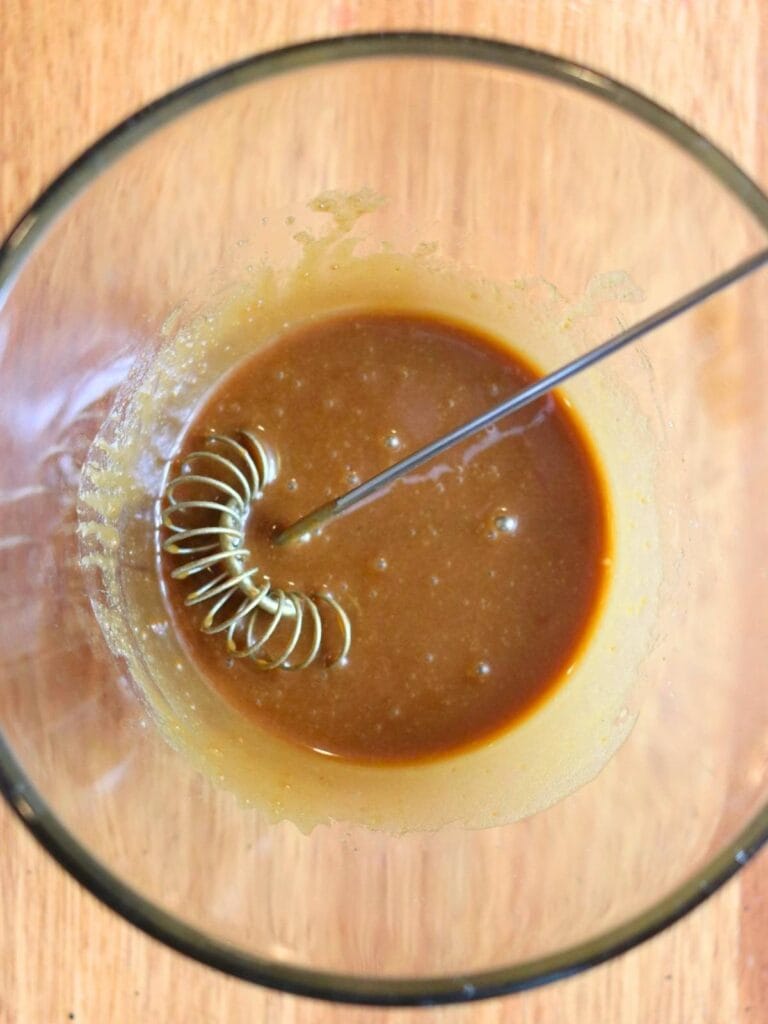 A glass bowl containing rich homemade maple glaze with a metal whisk immersed in it, placed on a wooden surface, conveys a cozy, homemade feel.