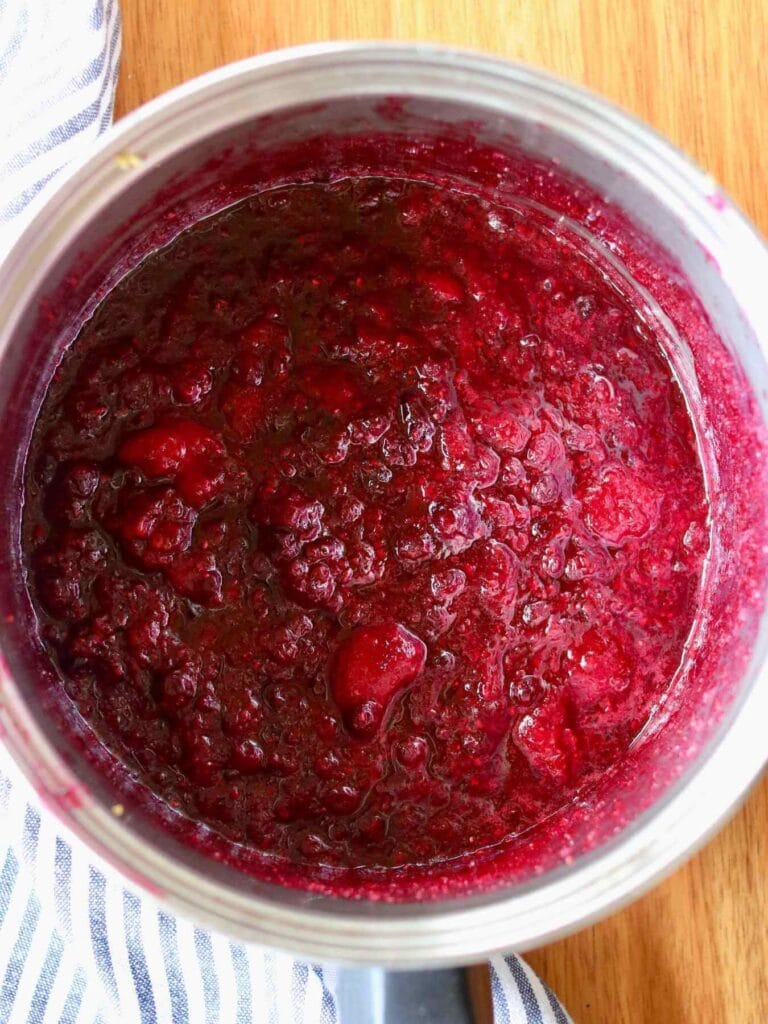 Rich, deep red mixed berry compote with a textured, chunky consistency fills a pot. The pot sits on a wooden table, partially covered by a striped cloth.