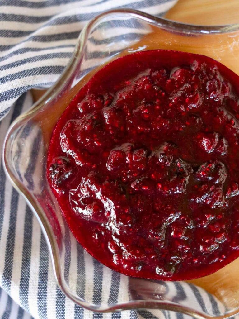 A close-up of vibrant red mixed berry compote in a glass dish, set on a wood surface beside a black-and-white striped cloth, conveying a festive and warm tone.