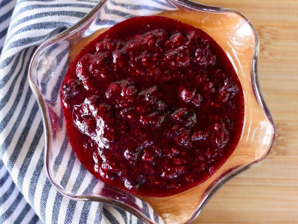 A glass bowl of vibrant red mixed berry compote sits on a wooden surface, next to a blue-striped cloth. The compote looks glossy and thick.