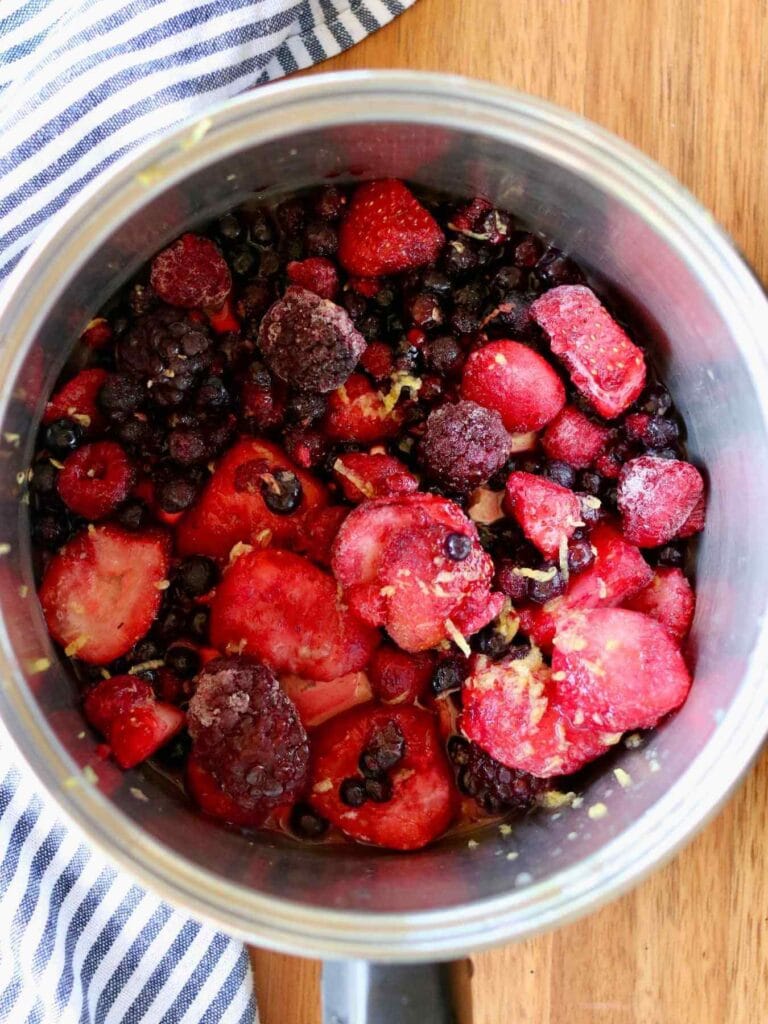 A pot filled with lemon juice, lemon zest, and frozen mixed berries, including strawberries, blueberries, and raspberries, sits on a wooden surface next to a striped cloth.