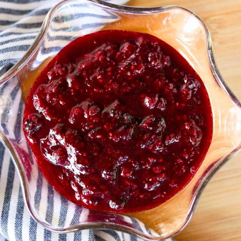 A glass bowl filled with rich, red mixed berry compote featuring a chunky texture. It rests on a wooden surface beside a striped cloth, evoking a cozy, homemade feel.