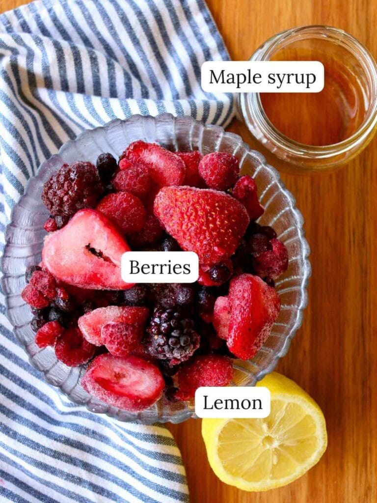 A glass bowl of frozen mixed berries on a striped cloth, next to a halved lemon and a jar of maple syrup on a wooden surface. Fresh and vibrant.