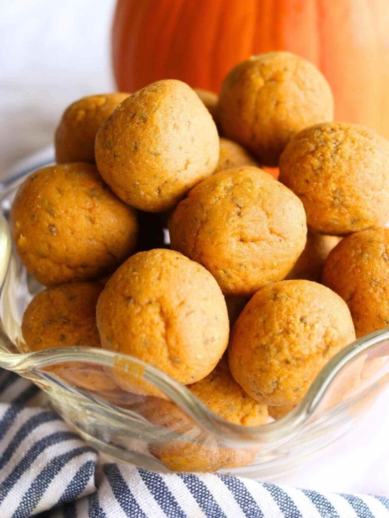 Glass bowl filled with round, textured pumpkin protein balls on a striped cloth. The backdrop includes a large, blurred orange pumpkin, creating a cozy autumn vibe.