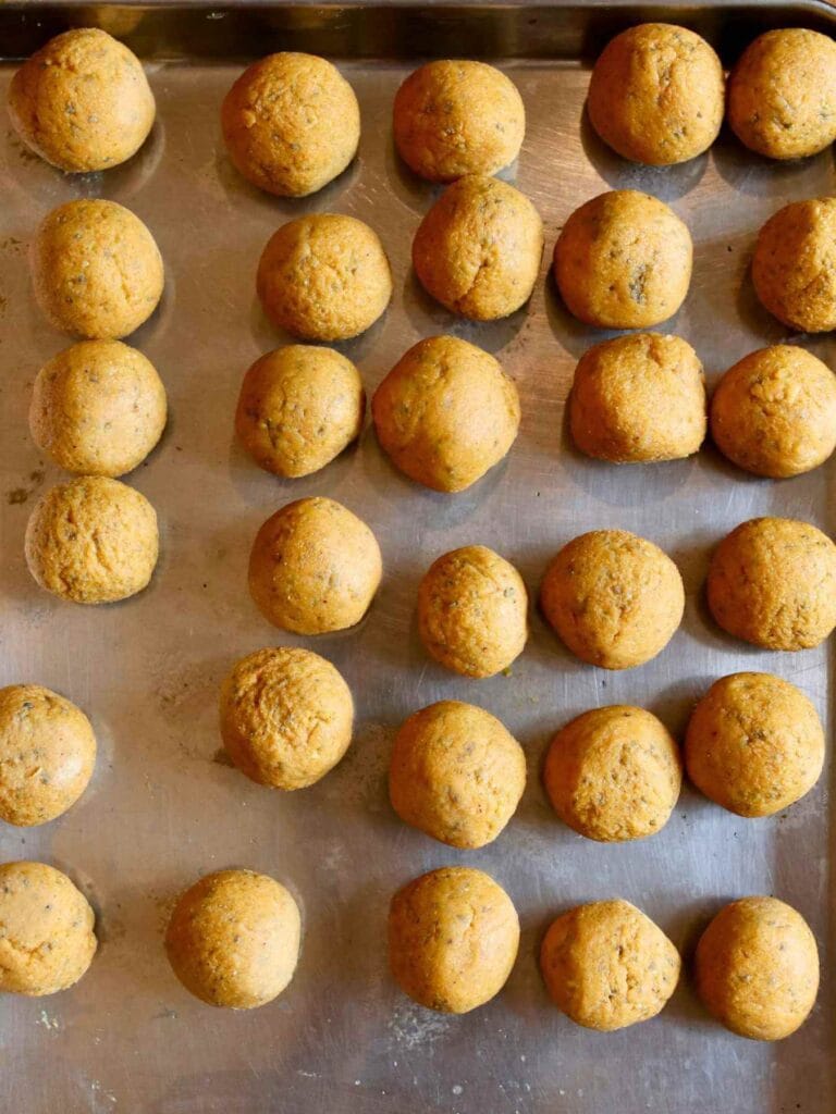 Round balls of pumpkin protein mixture arranged in neat rows on a metal baking tray. The balls are speckled and have a warm, golden-brown hue.