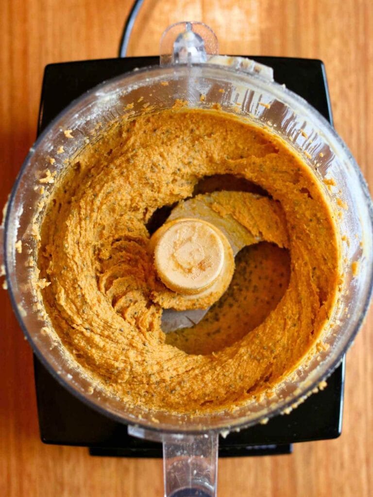 A food processor with creamy orange pumpkin mixture blended smoothly, seen from above. The wooden background adds a warm, homemade touch.