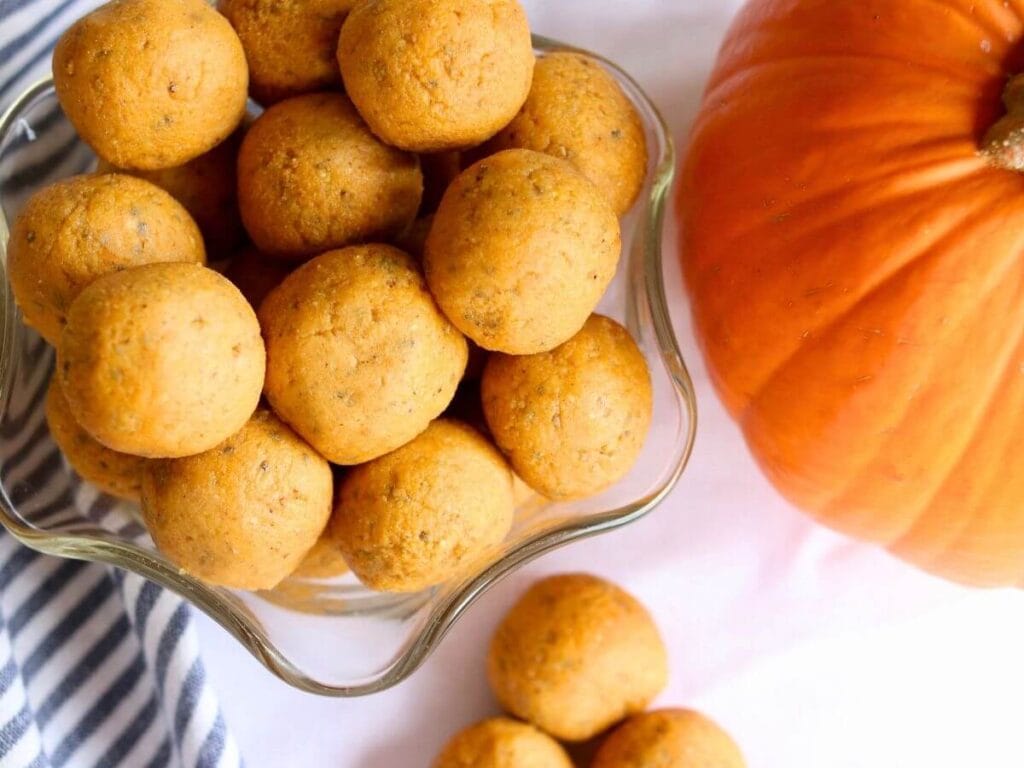 A bowl filled with round pumpkin protein balls, set on a white cloth with blue stripes. A bright orange pumpkin lies beside the bowl, creating a cozy, autumnal feel.