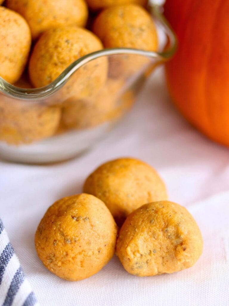 Three pumpkin protein balls on a white cloth, with more in a glass bowl in the background. The scene has a cozy, autumnal feel.