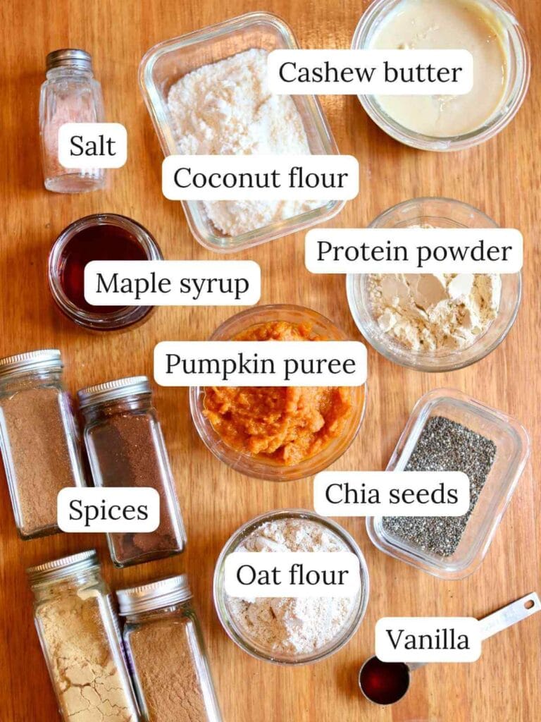Various ingredients, including cashew butter, oat flour, chia seeds, and pumpkin puree, are labeled on a wooden surface, suggesting a baking setup for pumpkin protein balls.