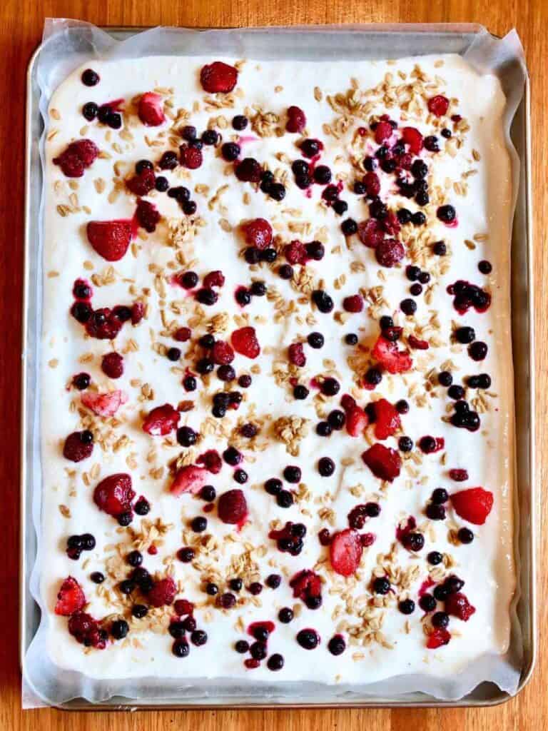 Close-up of a parchment-lined tray with yogurt bark ready for the freezer, topped with granola, strawberries, and blueberries. The texture is creamy and vibrant, evoking freshness.