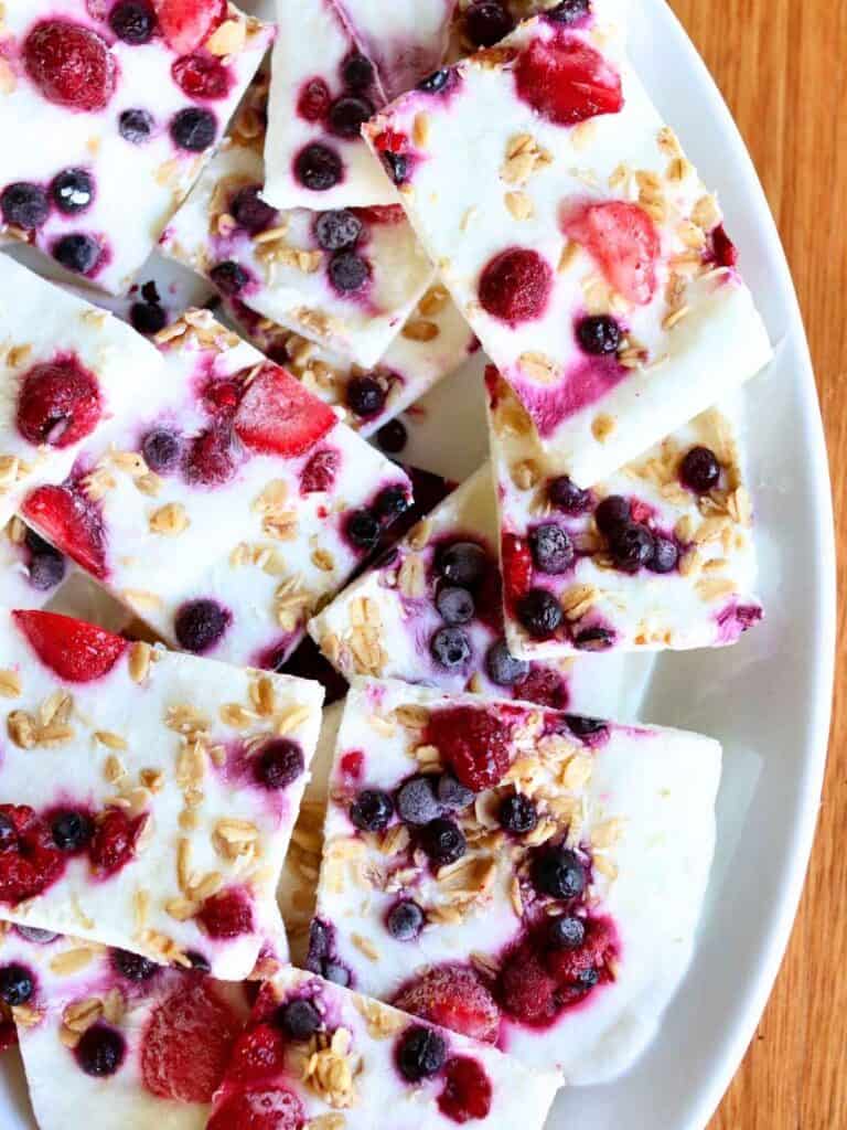 Frozen yogurt bark topped with vibrant red strawberries, blueberries, and granola. Presented on a white plate, offering a fresh, colorful snack.