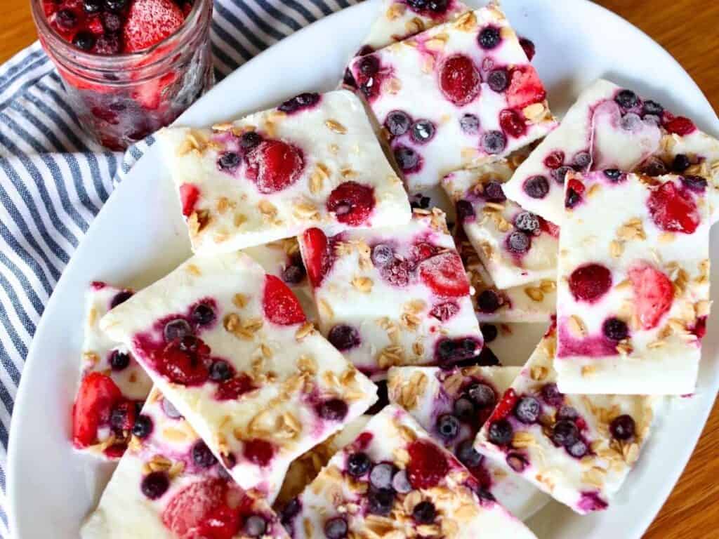 Frozen yogurt bark topped with mixed berries and granola, artfully arranged on a white plate. A jar of berries sits nearby on a striped cloth.