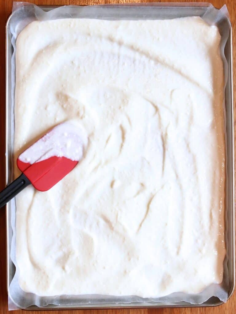 Creamy white yogurt evenly spread on a parchment-lined baking tray, with a red spatula gently smoothing the surface, creating a fresh, homemade cooking vibe.
