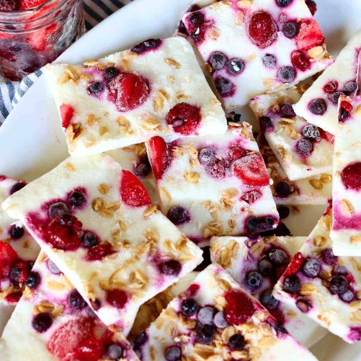 Close up of frozen yogurt bark topped with mixed berries and granola, arranged on a white plate. A jar of frozen berries sits nearby on a striped cloth.