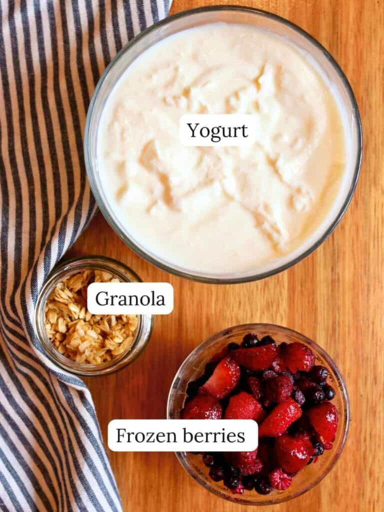 Aerial view of a wooden table with a bowl of creamy yogurt, a small glass of granola, and a bowl of frozen berries beside a striped cloth napkin.