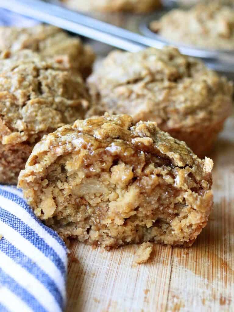 Half of a freshly baked moist apple oat muffin on a wooden surface with more muffins in the background.
