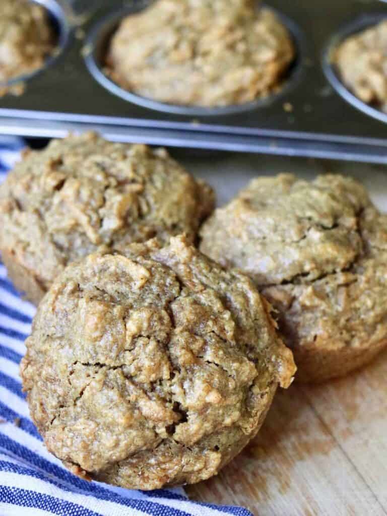 Freshly baked apple oat muffins in a muffin tin and on a wooden surface, next to a striped cloth napkin.